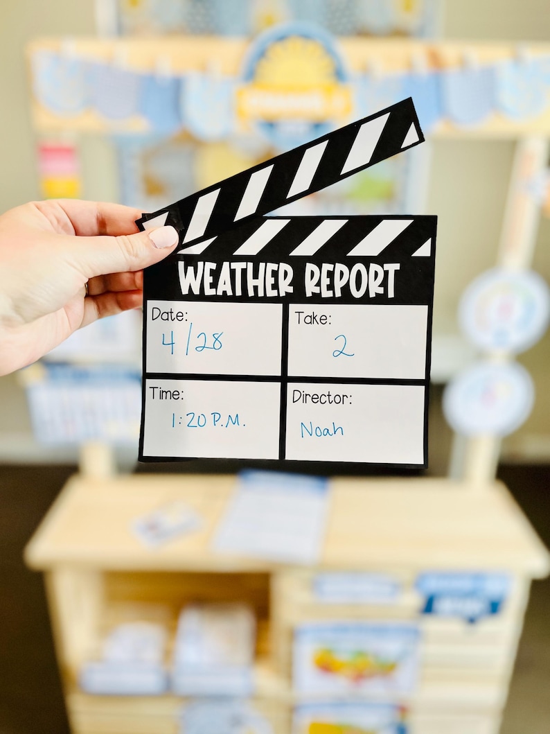 May include: A black and white clapperboard with the text "WEATHER REPORT" and handwritten details: date 4/28, take 2, time 1:20 P.M., and director Noah. The clapperboard is held in front of a wooden play stand.