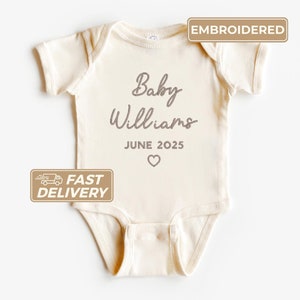 May include: A cream-colored baby onesie with the text "Baby Williams June 2025" embroidered in gray. The onesie has a small heart embroidered below the text. The onesie is labeled "Embroidered" and "Fast Delivery".