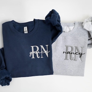 Peut inclure: Deux sweat-shirts ras du cou, un bleu marine et un gris. Le sweat-shirt bleu marine porte l'inscription "RN grace" en écriture blanche, et le sweat-shirt gris porte l'inscription "RN nancy" en écriture grise. Les deux sweat-shirts sont pliés.