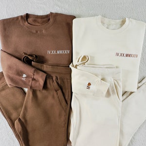 Puede incluir: Dos conjuntos a juego de pantalones de ch&aacute;ndal y sudaderas de color marr&oacute;n y crema. Las sudaderas tienen la fecha en n&uacute;meros romanos "IV.XX.MMXIV" impresa en la parte delantera. Los pantalones de ch&aacute;ndal tienen un peque&ntilde;o coraz&oacute;n con la letra "A" en un conjunto y la letra "M" en el otro.