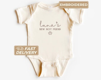 May include: A cream-colored baby onesie with short sleeves and a snap closure. The onesie has the text "Luna's New Best Friend" and a paw print embroidered in brown.