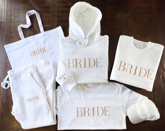 May include: A set of white bridal apparel with gold lettering. The set includes a hoodie, a sweatshirt, a t-shirt, sweatpants, a tote bag, and a baseball cap. All items feature the word "BRIDE" in gold lettering.