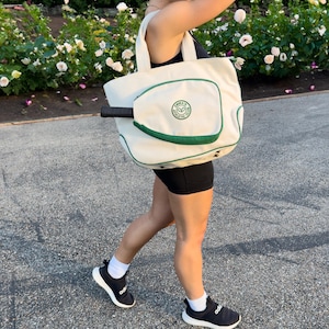 Peut inclure: Un sac fourre-tout en toile blanche avec une bordure verte et une poche pour raquette de tennis. Le sac porte un logo circulaire vert avec le texte "JAMAICA ATHLETIC CLUB".