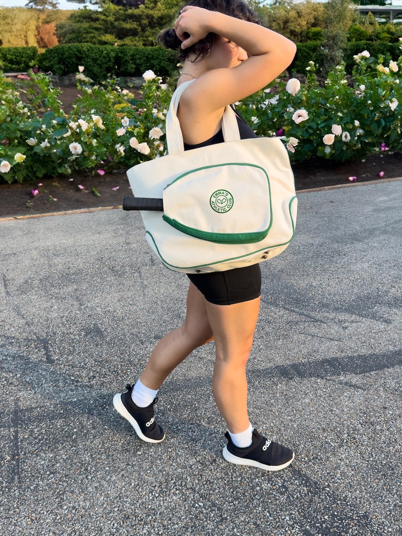 Peut inclure: Un sac fourre-tout en toile blanche avec une bordure verte et une poche pour raquette de tennis. Le sac porte un logo circulaire vert avec le texte "JAMAICA ATHLETIC CLUB".