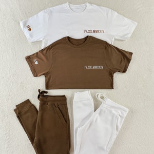 Puede incluir: Dos camisetas blancas con texto marr&oacute;n que dice "IV.XX.MMXIV" y una camiseta marr&oacute;n con texto blanco que dice "IV.XX.MMXIV". La camiseta marr&oacute;n tiene un coraz&oacute;n blanco con la letra "A" dentro. Tambi&eacute;n hay dos pares de pantalones de ch&aacute;ndal, uno marr&oacute;n y uno blanco.