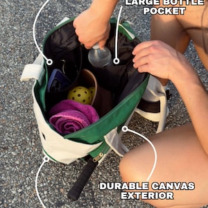 Peut inclure: Un sac fourre-tout en toile vert et blanc avec une poche int&eacute;rieure zipp&eacute;e, une grande poche pour bouteille et des pieds en laiton. Le sac est rempli de divers articles, dont une bouteille d'eau, une serviette rose et quelques balles.