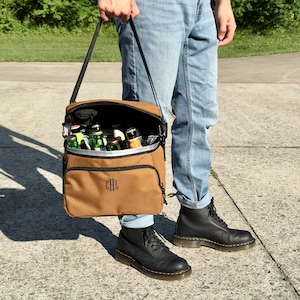 Pu&ograve; includere: Una borsa termica in tela marrone con una tracolla nera e una cerniera nera. La borsa &egrave; piena di bottiglie di birra e ha le iniziali "EHL" ricamate sul davanti.