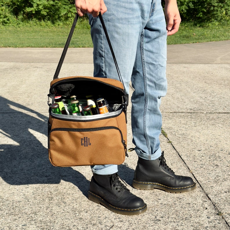 Pu&ograve; includere: Una borsa termica in tela marrone con una tracolla nera e una cerniera nera. La borsa &egrave; piena di bottiglie di birra e ha le iniziali "EHL" ricamate sul davanti.