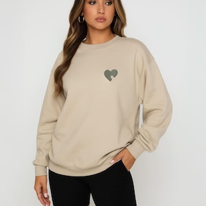 K&ouml;nnte beinhalten: Beigefarbenes Sweatshirt mit einem gr&uuml;nen herzf&ouml;rmigen Aufdruck auf der Brust. Das Sweatshirt wird mit schwarzen Shorts kombiniert. Die Person tr&auml;gt goldene Creolen.