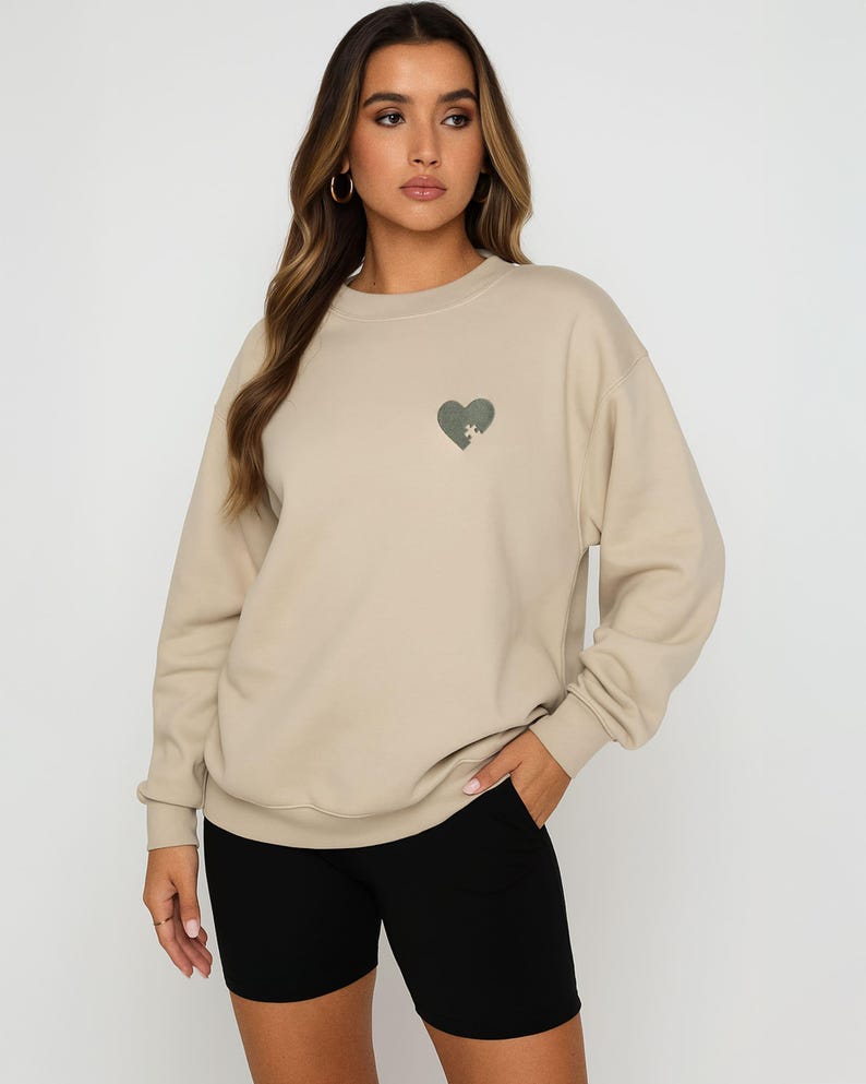 K&ouml;nnte beinhalten: Beigefarbenes Sweatshirt mit einem gr&uuml;nen herzf&ouml;rmigen Aufdruck auf der Brust. Das Sweatshirt wird mit schwarzen Shorts kombiniert. Die Person tr&auml;gt goldene Creolen.