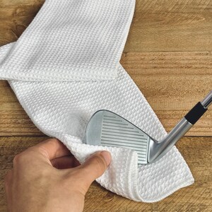Peut inclure: Une serviette de nettoyage de club de golf en microfibre blanche avec un fer &agrave; repasser de club de golf argent&eacute; pos&eacute; dessus.