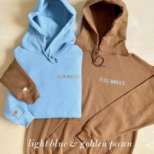K&ouml;nnte beinhalten: Zwei Kapuzenpullover, einer hellblau und einer goldbraun, werden gezeigt. Jeder hat eine Kapuze mit Kordelzug und eine Vordertasche. Der hellblaue Pullover hat einen braunen &Auml;rmel mit gestickten Initialen und einem Herz. Beide haben das Datum IV.XX.MMXXIV.