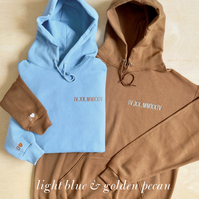 K&ouml;nnte beinhalten: Zwei Kapuzenpullover, einer hellblau und einer goldbraun, werden gezeigt. Jeder hat eine Kapuze mit Kordelzug und eine Vordertasche. Der hellblaue Pullover hat einen braunen &Auml;rmel mit gestickten Initialen und einem Herz. Beide haben das Datum IV.XX.MMXXIV.
