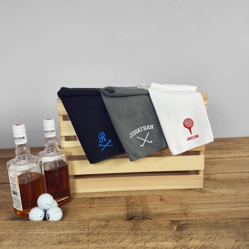 Peut inclure: Trois serviettes de golf en tissu gaufr&eacute; avec broderie personnalis&eacute;e. Les serviettes sont en bleu marine, gris et blanc. La serviette bleu marine a un "R" bleu avec des clubs de golf crois&eacute;s. La serviette grise porte le nom "JONATHAN" avec des clubs de golf crois&eacute;s. La serviette blanche a une balle de golf rouge avec le nom "JACOB".