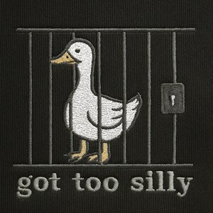 K&ouml;nnte beinhalten: Schwarzer Stoff mit einem gestickten Design, das eine wei&szlig;e Ente in einem K&auml;fig zeigt. Die Ente hat einen gelben Schnabel und gelbe F&uuml;&szlig;e. Der Text "got too silly" ist unter dem K&auml;fig gestickt.