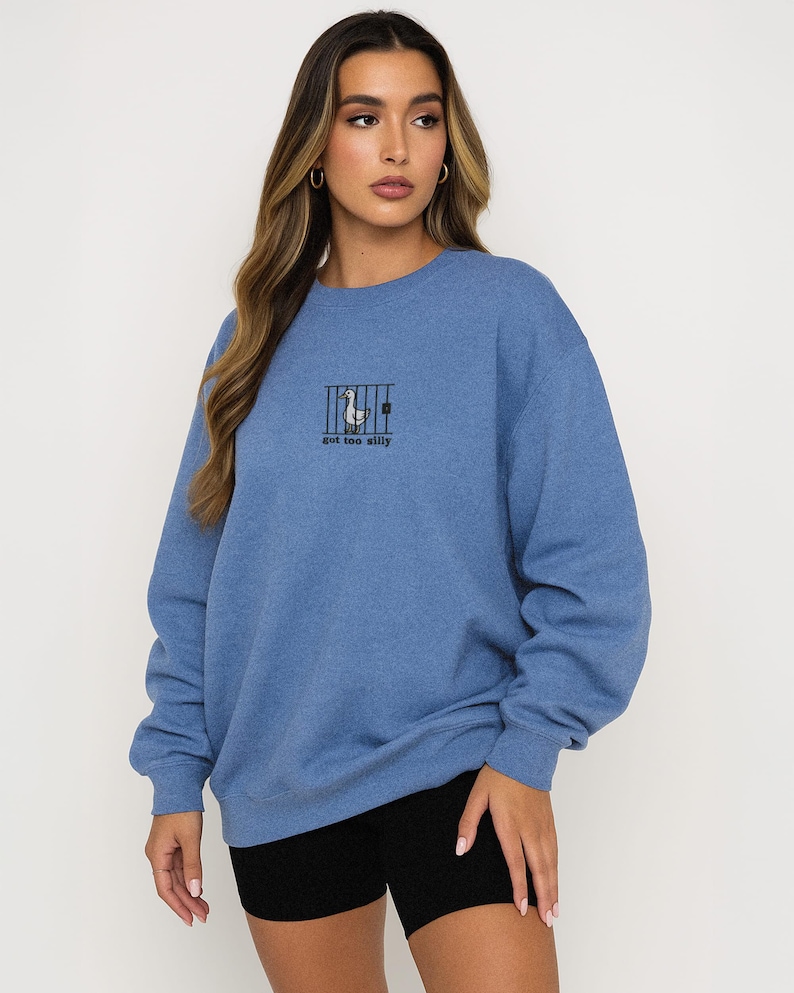 K&ouml;nnte beinhalten: Blaues Sweatshirt mit einem Enten-Grafik hinter Gittern und dem Text "got too silly". Das Sweatshirt hat eine blaue Farbe und lange &Auml;rmel. Das Model tr&auml;gt schwarze Shorts.