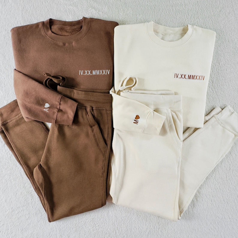 Puede incluir: Dos conjuntos a juego de pantalones de ch&aacute;ndal y sudaderas de color marr&oacute;n y crema. Las sudaderas tienen la fecha en n&uacute;meros romanos "IV.XX.MMXIV" impresa en la parte delantera. Los pantalones de ch&aacute;ndal tienen un peque&ntilde;o coraz&oacute;n con la letra "A" en un conjunto y la letra "M" en el otro.