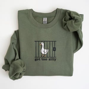 K&ouml;nnte beinhalten: Olivgr&uuml;nes Sweatshirt mit Rundhalsausschnitt. Auf der Vorderseite befindet sich ein gesticktes Design einer wei&szlig;en Ente hinter Gittern mit dem Text "got too silly" in Schwarz. Die &Auml;rmel sind gefaltet.