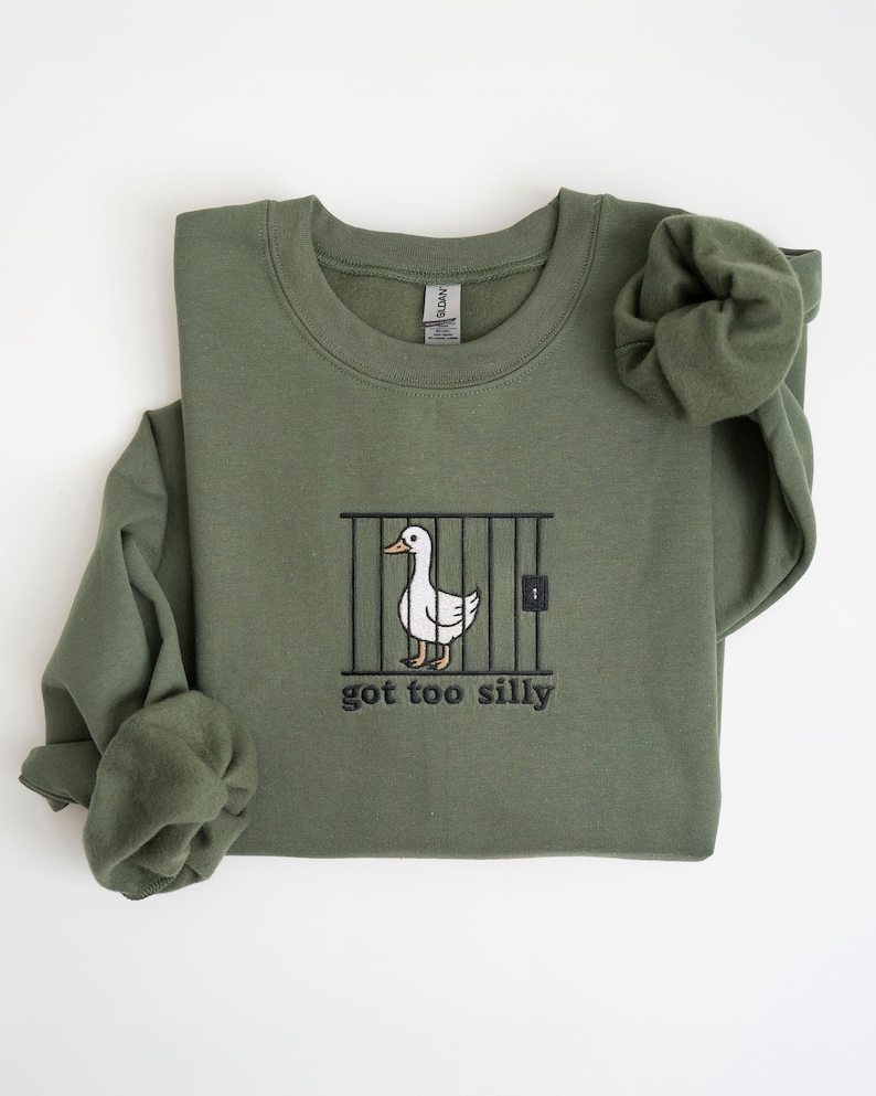 K&ouml;nnte beinhalten: Olivgr&uuml;nes Sweatshirt mit Rundhalsausschnitt. Auf der Vorderseite befindet sich ein gesticktes Design einer wei&szlig;en Ente hinter Gittern mit dem Text "got too silly" in Schwarz. Die &Auml;rmel sind gefaltet.