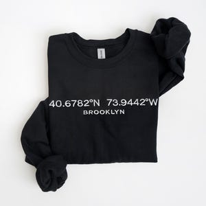May include: Black crewneck sweatshirt with white text that reads "40.6782°N 73.9442°W BROOKLYN". The sweatshirt is folded and has long sleeves. The Gildan brand tag is visible.