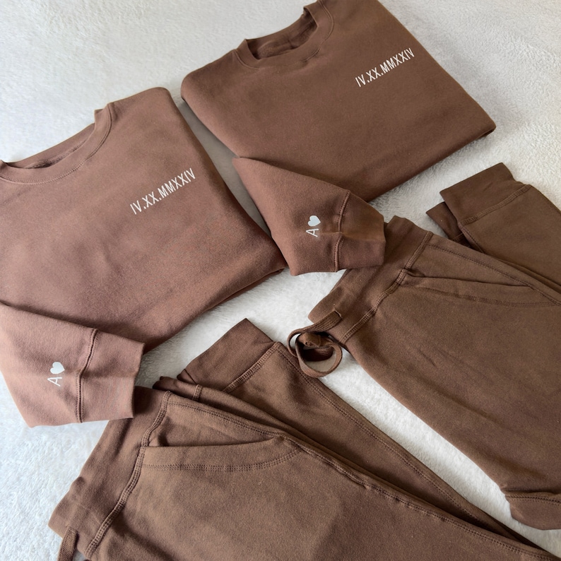 Puede incluir: Dos sudaderas marrones con texto bordado en blanco, "IV.XX.MMXIV", y un peque&ntilde;o coraz&oacute;n con la letra "A" en la manga. Dos pares de pantalones de ch&aacute;ndal marrones con cord&oacute;n.