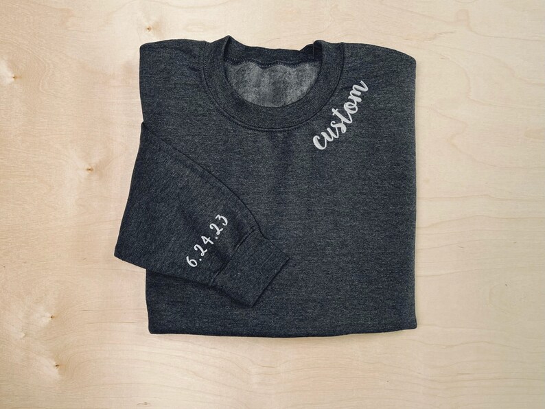 Op de afbeelding: Een donkergrijs sweatshirt met het woord "custom" geborduurd op de voorkant en de datum "6.24.23" geborduurd op de mouw.