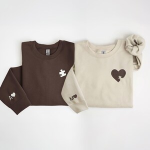 K&ouml;nnte beinhalten: Zwei Sweatshirts, eines braun und eines beige, werden gezeigt. Das braune Sweatshirt hat ein wei&szlig;es Puzzleteil auf der Brust und ein "A" mit einem Herz auf dem &Auml;rmel. Das beige Sweatshirt hat ein Herz mit einem Puzzleteil und ein "M" mit einem Herz.