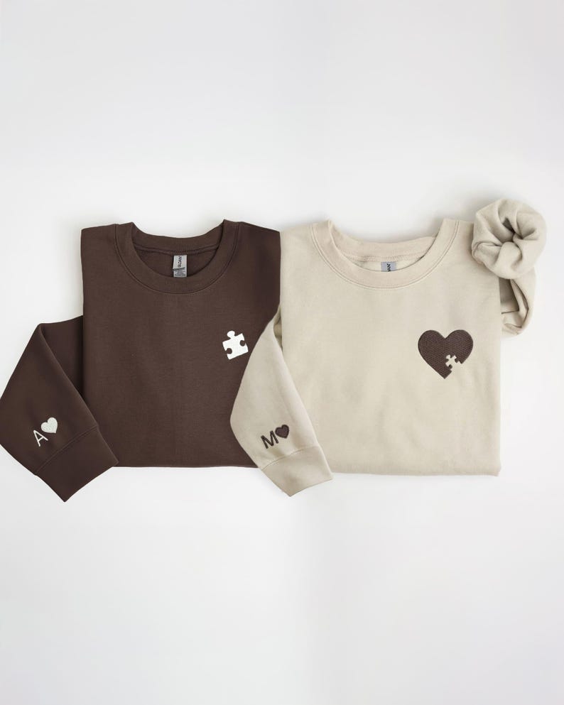 K&ouml;nnte beinhalten: Zwei Sweatshirts, eines braun und eines beige, werden gezeigt. Das braune Sweatshirt hat ein wei&szlig;es Puzzleteil auf der Brust und ein "A" mit einem Herz auf dem &Auml;rmel. Das beige Sweatshirt hat ein Herz mit einem Puzzleteil und ein "M" mit einem Herz.