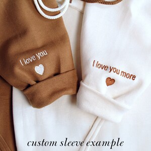 K&ouml;nnte beinhalten: Nahaufnahme von zwei lang&auml;rmeligen Kleidungsst&uuml;cken, eines braun und eines wei&szlig;. Der braune &Auml;rmel hat "I love you" in Wei&szlig; mit einem wei&szlig;en Herz gestickt. Der wei&szlig;e &Auml;rmel hat "I love you more" in Braun mit einem braunen Herz gestickt. Der Text unten lautet "custom sleeve example".