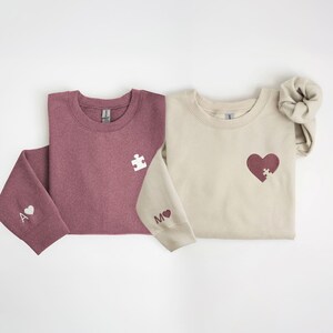 K&ouml;nnte beinhalten: Zwei Sweatshirts mit Rundhalsausschnitt, eines in Burgunderrot und eines in Beige, mit passenden Puzzleteilen und Herzmotiven. Das burgunderfarbene Sweatshirt hat ein wei&szlig;es Puzzleteil und ein Herz mit dem Buchstaben "A" auf dem &Auml;rmel. Das beige Sweatshirt hat ein Herz mit einem Puzzleteil und dem Buchstaben "M" auf dem &Auml;rmel.