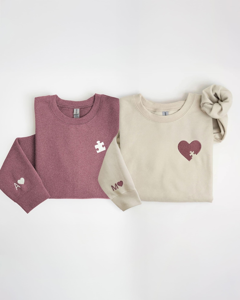 K&ouml;nnte beinhalten: Zwei Sweatshirts mit Rundhalsausschnitt, eines in Burgunderrot und eines in Beige, mit passenden Puzzleteilen und Herzmotiven. Das burgunderfarbene Sweatshirt hat ein wei&szlig;es Puzzleteil und ein Herz mit dem Buchstaben "A" auf dem &Auml;rmel. Das beige Sweatshirt hat ein Herz mit einem Puzzleteil und dem Buchstaben "M" auf dem &Auml;rmel.
