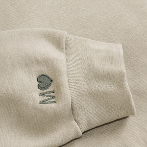 K&ouml;nnte beinhalten: Nahaufnahme eines beigefarbenen Sweatshirt-&Auml;rmels. Das B&uuml;ndchen ist gerippt, und der &Auml;rmel zeigt ein gesticktes Design eines Herzens &uuml;ber dem Buchstaben "M" in einem gedeckten Gr&uuml;n. Der Stoff hat ein weiches, strukturiertes Aussehen.