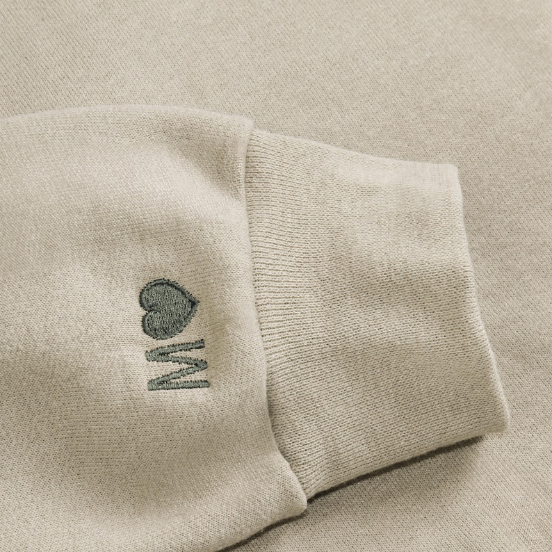 K&ouml;nnte beinhalten: Nahaufnahme eines beigefarbenen Sweatshirt-&Auml;rmels. Das B&uuml;ndchen ist gerippt, und der &Auml;rmel zeigt ein gesticktes Design eines Herzens &uuml;ber dem Buchstaben "M" in einem gedeckten Gr&uuml;n. Der Stoff hat ein weiches, strukturiertes Aussehen.