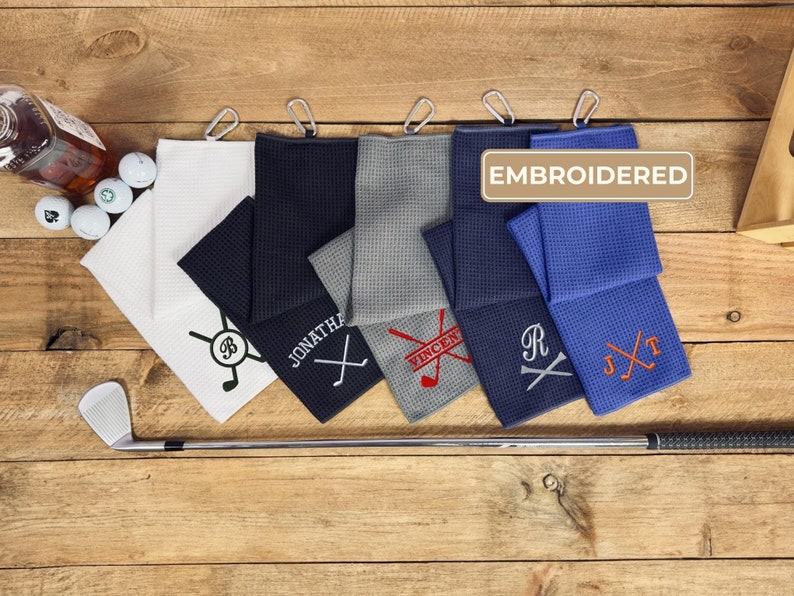 Peut inclure: Cinq serviettes de golf brod&eacute;es de diff&eacute;rentes couleurs, dont blanc, noir, gris, bleu et bleu marine. Chaque serviette a un motif brod&eacute; diff&eacute;rent, y compris un club de golf et des initiales. Les serviettes sont accroch&eacute;es sur une surface en bois avec une bouteille de whisky et des balles de golf en arri&egrave;re-plan. Le texte "EMBROIDERED" est visible dans le coin sup&eacute;rieur droit de l'image.