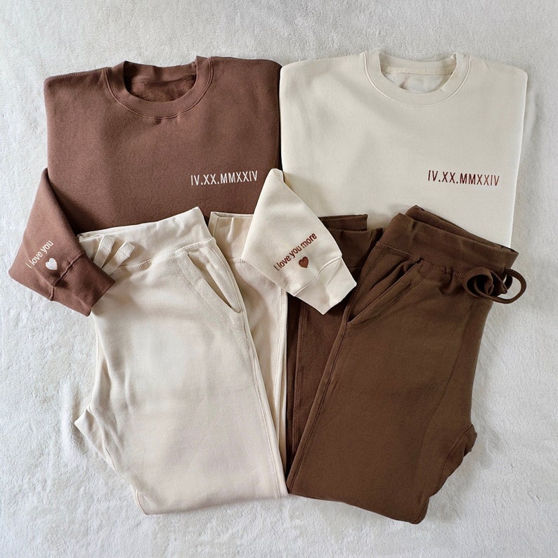 Puede incluir: Dos conjuntos a juego de pantalones de ch&aacute;ndal y sudaderas de color marr&oacute;n y crema. Las sudaderas tienen el texto "IV.XX.MMXIV" en la parte delantera y los pantalones de ch&aacute;ndal tienen el texto "I love you" y "I love you more" en los pu&ntilde;os.