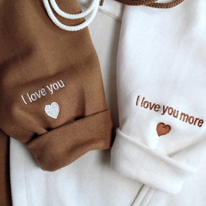 Engagement gift for couple shown. I love you / te amo embroidered with heart on sleeve on left. I love you more / te amo mas example shown on sleeve on right. Custom initial and text or phrase or wording on sleeve.