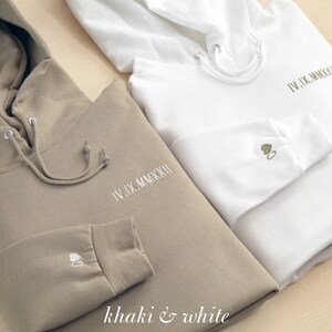 K&ouml;nnte beinhalten: Zwei Kapuzenpullover, einer in Khaki und einer in Wei&szlig;, werden gezeigt. Beide haben gestickten Text und ein kleines Herzdesign auf dem &Auml;rmel. Der khakifarbene Pullover hat wei&szlig;e Stickereien, w&auml;hrend der wei&szlig;e olivgr&uuml;ne Stickereien hat. Der Text "IV.IX.MMXXII" ist auf der Brust gestickt.