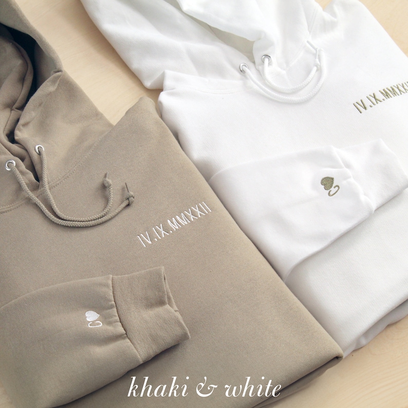K&ouml;nnte beinhalten: Zwei Kapuzenpullover, einer in Khaki und einer in Wei&szlig;, werden gezeigt. Beide haben gestickten Text und ein kleines Herzdesign auf dem &Auml;rmel. Der khakifarbene Pullover hat wei&szlig;e Stickereien, w&auml;hrend der wei&szlig;e olivgr&uuml;ne Stickereien hat. Der Text "IV.IX.MMXXII" ist auf der Brust gestickt.