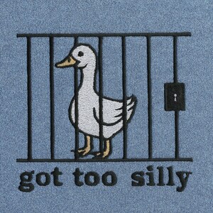 K&ouml;nnte beinhalten: Hellblaues Sweatshirt mit einer Stickerei eines wei&szlig;en Entchens in einem schwarzen K&auml;fig. Das Entchen hat einen gelben Schnabel und gelbe F&uuml;&szlig;e. Der Text "got too silly" ist unter dem K&auml;fig in Schwarz gestickt.