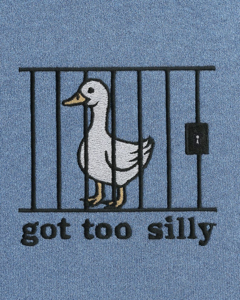 K&ouml;nnte beinhalten: Hellblaues Sweatshirt mit einer Stickerei eines wei&szlig;en Entchens in einem schwarzen K&auml;fig. Das Entchen hat einen gelben Schnabel und gelbe F&uuml;&szlig;e. Der Text "got too silly" ist unter dem K&auml;fig in Schwarz gestickt.