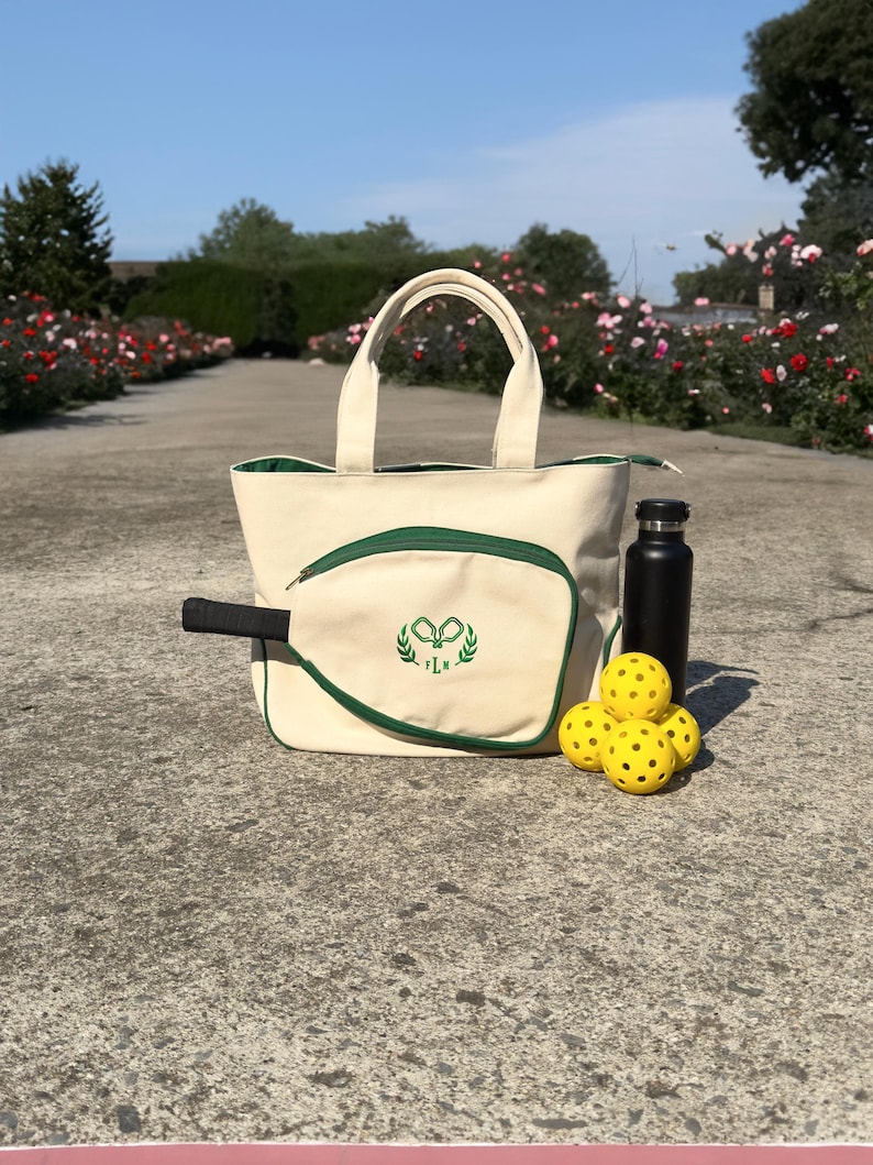 Peut inclure: Un sac fourre-tout en toile blanche avec une bordure verte et un logo brod&eacute; vert de deux raquettes crois&eacute;es et une couronne de laurier. Le sac est rempli de balles de pickleball jaunes et d'une bouteille d'eau noire. Le sac est pos&eacute; sur une surface en b&eacute;ton gris.