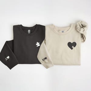 K&ouml;nnte beinhalten: Zwei Sweatshirts, eines in Schwarz und eines in Beige, werden gezeigt. Das schwarze Sweatshirt hat ein wei&szlig;es Puzzleteil auf der Brust und ein "A" mit einem Herz auf dem &Auml;rmel. Das beige Sweatshirt hat ein schwarzes Herz mit einem Puzzleteil und ein "M" mit einem Herz auf dem &Auml;rmel.