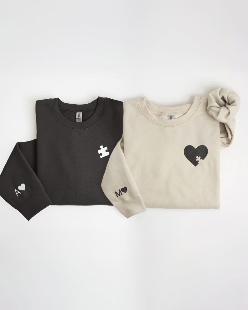 K&ouml;nnte beinhalten: Zwei Sweatshirts, eines in Schwarz und eines in Beige, werden gezeigt. Das schwarze Sweatshirt hat ein wei&szlig;es Puzzleteil auf der Brust und ein "A" mit einem Herz auf dem &Auml;rmel. Das beige Sweatshirt hat ein schwarzes Herz mit einem Puzzleteil und ein "M" mit einem Herz auf dem &Auml;rmel.