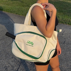 Peut inclure: Un sac fourre-tout en toile blanche avec une bordure verte et une poche s&eacute;par&eacute;e pour une raquette de pickleball. Le sac porte le texte "Lilly Pickleballer" brod&eacute; sur le devant.
