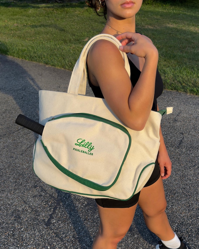 Peut inclure: Un sac fourre-tout en toile blanche avec une bordure verte et une poche s&eacute;par&eacute;e pour une raquette de pickleball. Le sac porte le texte "Lilly Pickleballer" brod&eacute; sur le devant.