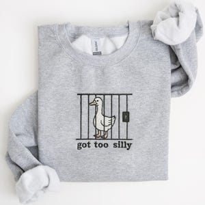 K&ouml;nnte beinhalten: Ein hellgraues Sweatshirt mit einem wei&szlig;en Enten-Motiv hinter schwarzen St&auml;ben und dem Text "got too silly" in Schwarz. Das Sweatshirt hat einen Rundhalsausschnitt und lange &Auml;rmel.