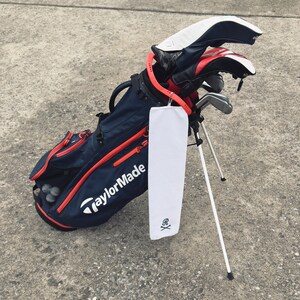 Peut inclure: Un sac de golf bleu et rouge avec une serviette blanche suspendue. La serviette a un motif de cr&acirc;ne et d'os crois&eacute;s avec la lettre "R" au centre. Le sac est sur un support blanc et porte le logo TaylorMade sur le c&ocirc;t&eacute;.