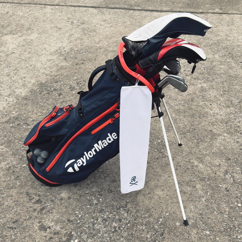 Peut inclure: Un sac de golf bleu et rouge avec une serviette blanche suspendue. La serviette a un motif de cr&acirc;ne et d'os crois&eacute;s avec la lettre "R" au centre. Le sac est sur un support blanc et porte le logo TaylorMade sur le c&ocirc;t&eacute;.