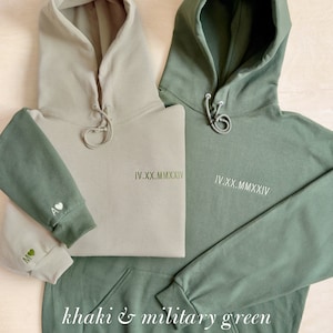K&ouml;nnte beinhalten: Zwei Kapuzenpullover in Khaki und Milit&auml;rgr&uuml;n. Jeder hat eine Kapuze mit Kordelzug und eine Vordertasche. Der khakifarbene Pullover hat einen milit&auml;rgr&uuml;nen &Auml;rmel, bestickt mit Initialen und einem Herz. Der milit&auml;rgr&uuml;ne Pullover hat das Datum "IV.XX.MMXXIV" auf der Brust gestickt.