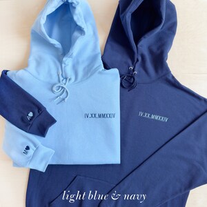 K&ouml;nnte beinhalten: Zwei Kapuzenpullover, einer hellblau und einer marineblau. Der hellblaue Pullover hat einen marineblauen &Auml;rmel mit Initialen und einem Herz. Beide Pullover haben das r&ouml;mische Datum "IV.XX.MMXXIV" auf der Vorderseite. Der Text "light blue & navy" steht unten.